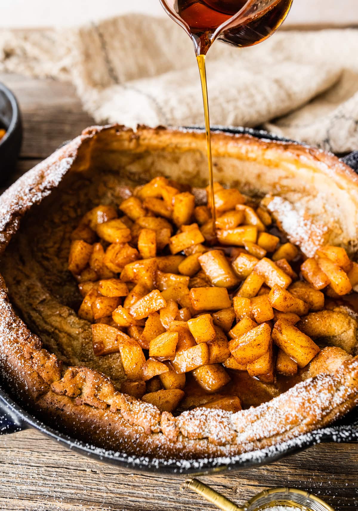 maple syrup being poured over brown butter Dutch baby with cinnamon apples in cast iron skillet. 